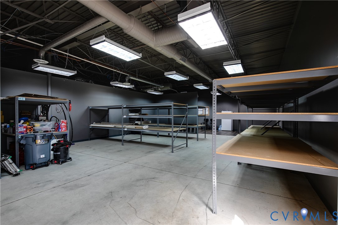 Industrial storage area featuring high-contrast dark walls, exposed ceiling ductwork, concrete flooring, and multiple overhead LED light fixtures