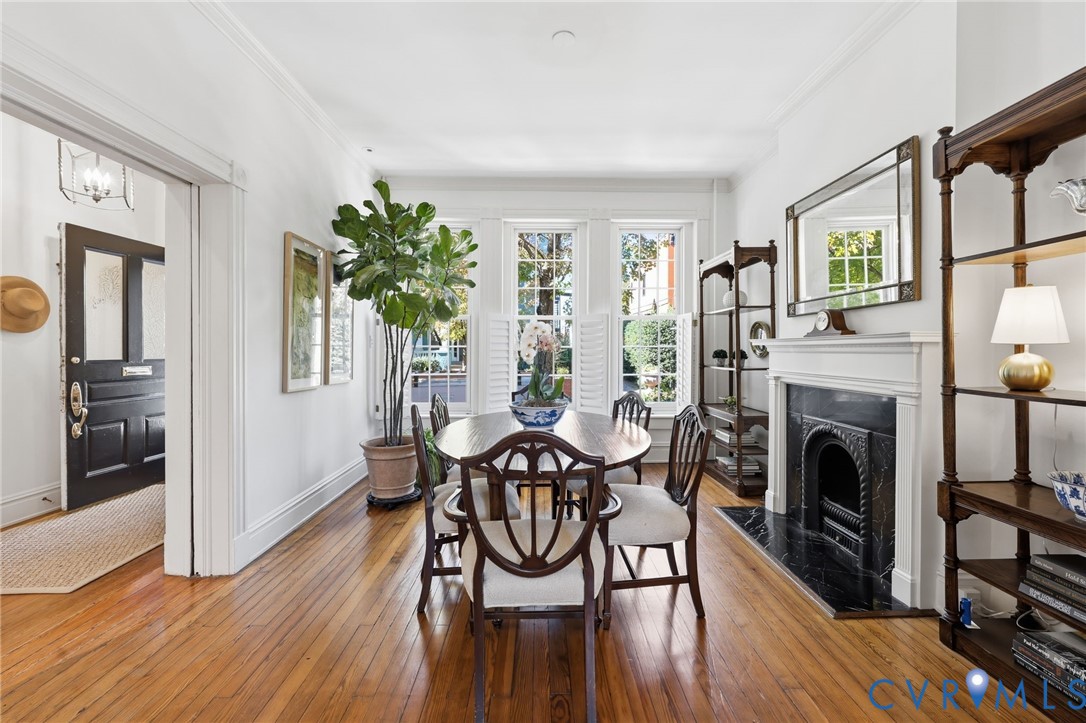 Hardwood flooring, triple-window configuration with plantation shutters, decorative fireplace featuring a black stone surround and a carved mantel, crown molding, and a dark-finished solid wood entry door with glass pane detail Hardwood flooring, triple-window configuration with plantation shutters, decorative fireplace featuring a black stone surround and a carved mantel, crown molding, and a dark-finished solid wood entry door with glass pane detail