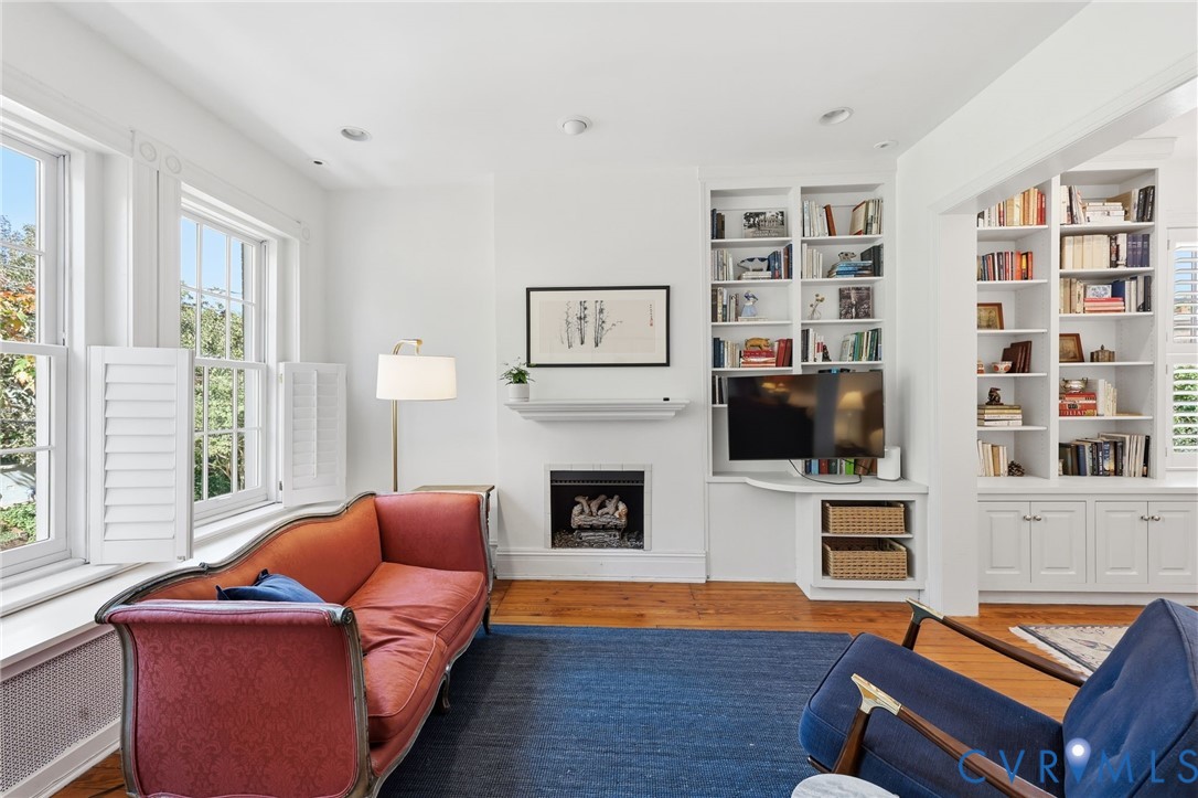 Fireplace with a mantel, built-in floor-to-ceiling shelving, wood-finish flooring, and multi-pane windows with plantation-style shutters