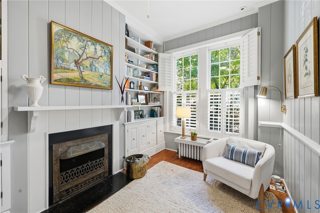 Fireplace with a decorative metal surround and hearth, vertical wood paneling, built-in floor-to-ceiling shelving, and windows featuring plantation shutters