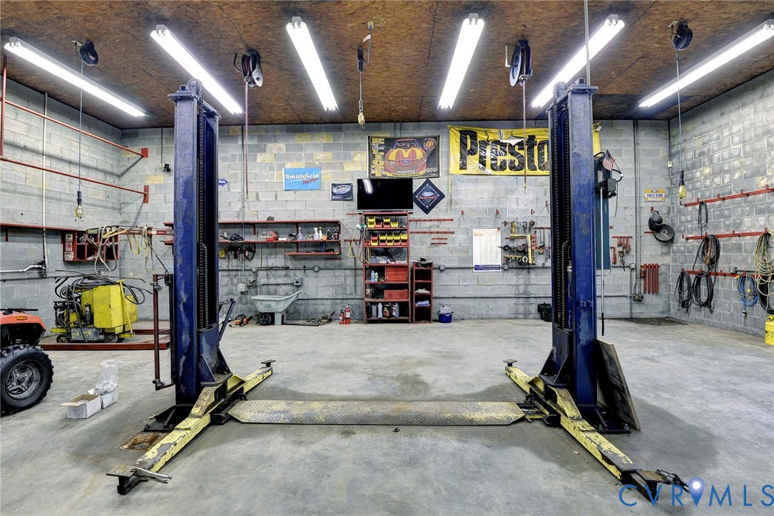 Industrial-style workshop featuring a concrete floor, two-post vehicle lift, exposed concrete block walls, overhead fluorescent lighting, and wall-mounted storage shelving