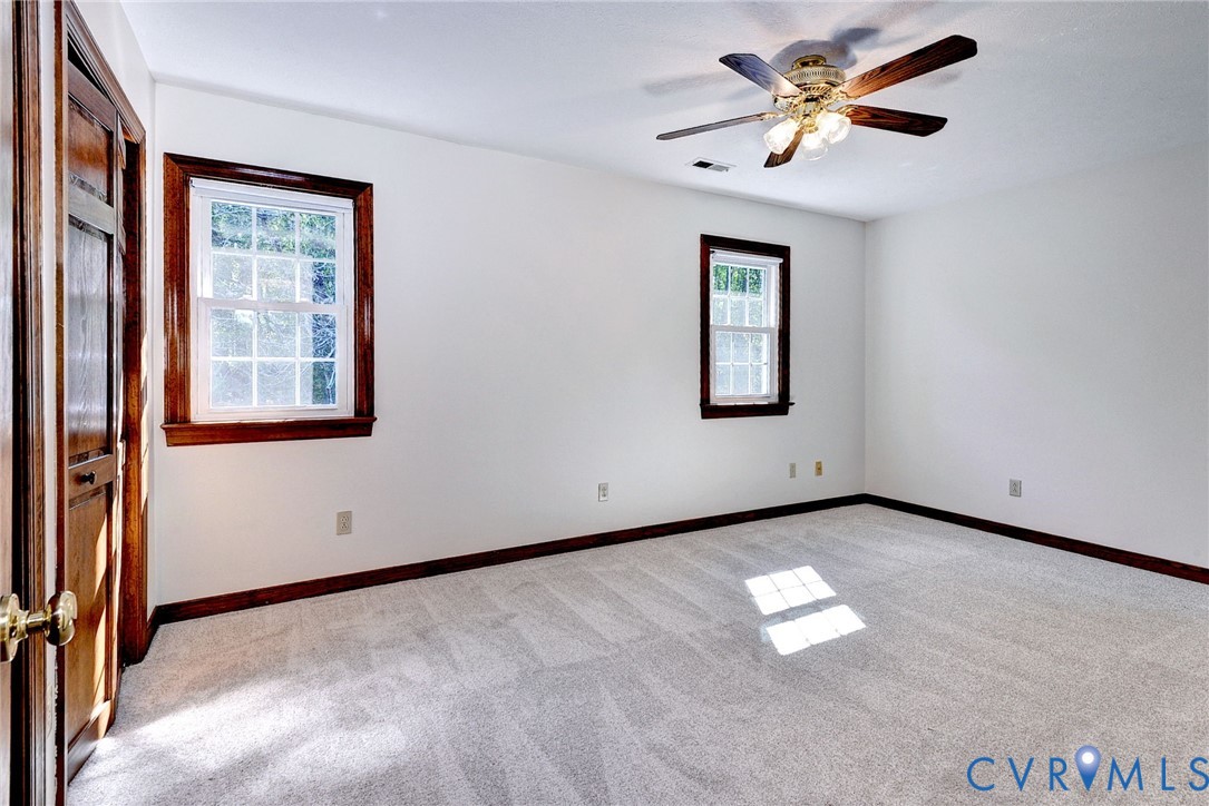 Neutral interior space featuring carpeted flooring, two multi-pane windows with wood-finish trim, a wood-finish interior door, and a ceiling-mounted light fixture with fan blades