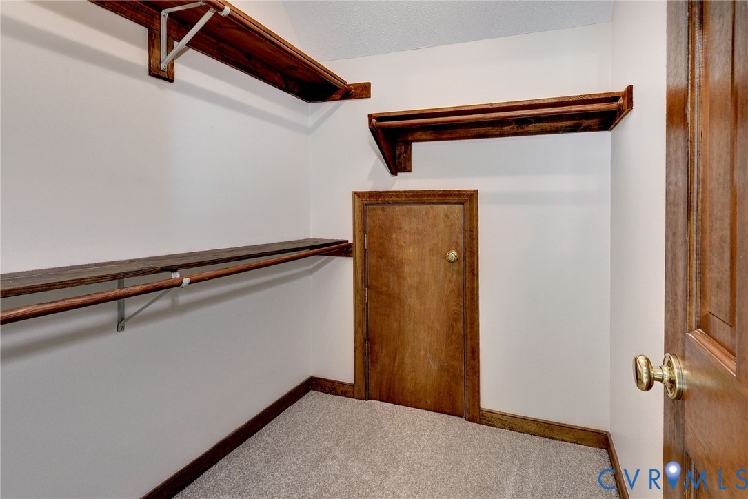 Walk-in storage space featuring built-in wood shelving, integrated garment rods, neutral carpeting, and a dedicated access door with a metallic knob