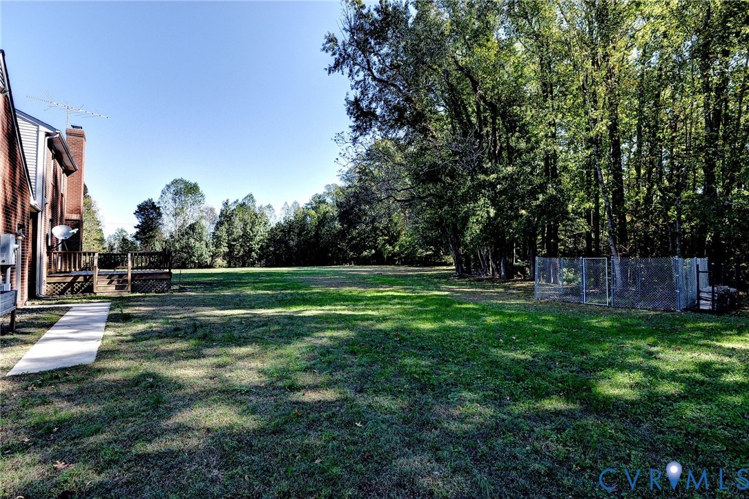 Expansive backyard featuring a concrete walkway, elevated wood deck, and perimeter chain-link fencing, bordered by mature trees