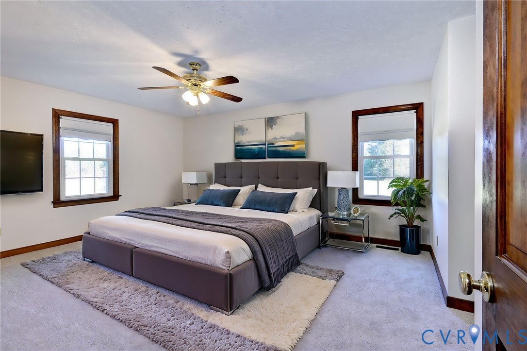 VIRTUAL STAGING - Spacious room featuring neutral carpeting, two windows with stained-wood trim, and a multi-blade ceiling fan with integrated lighting