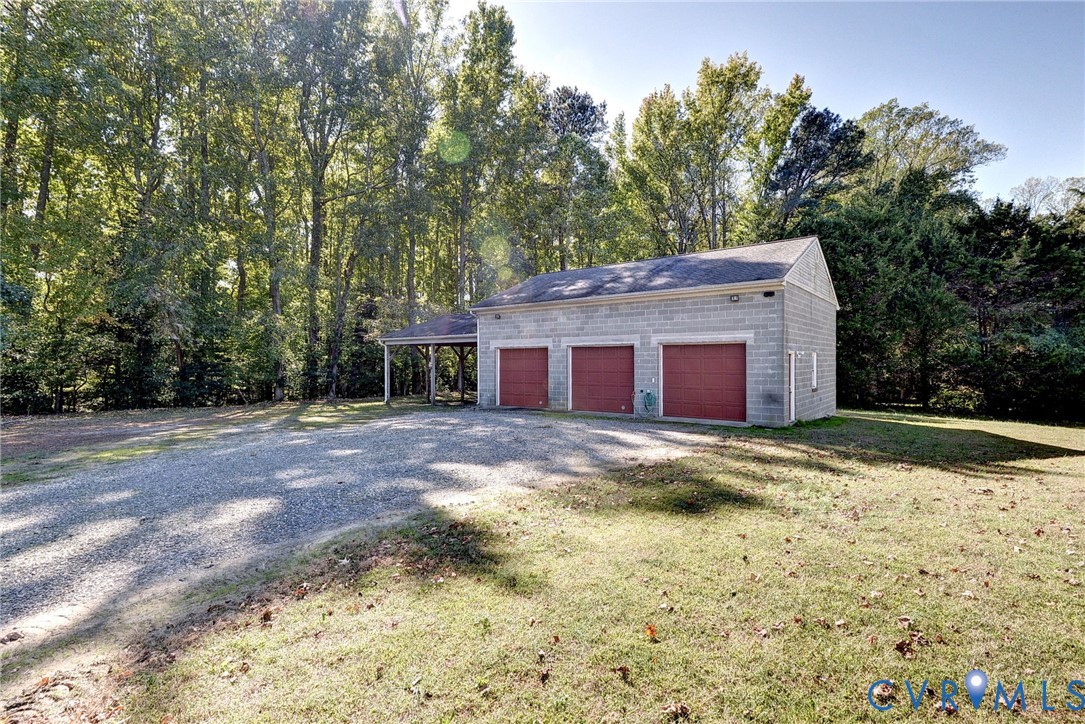 Three-car detached garage featuring masonry exterior, shingle roof, integrated side carport, and gravel driveway