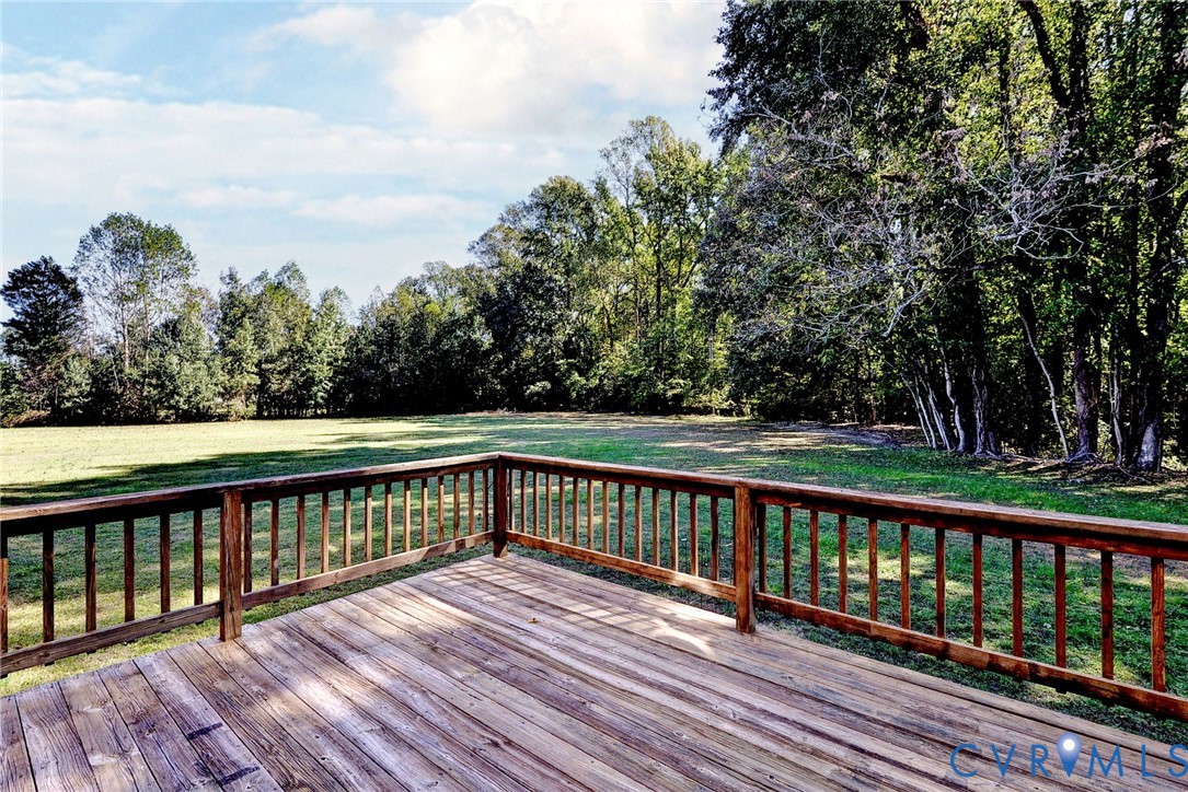 Elevated wooden deck featuring a perimeter railing, overlooking a sprawling grass lawn and a dense tree line