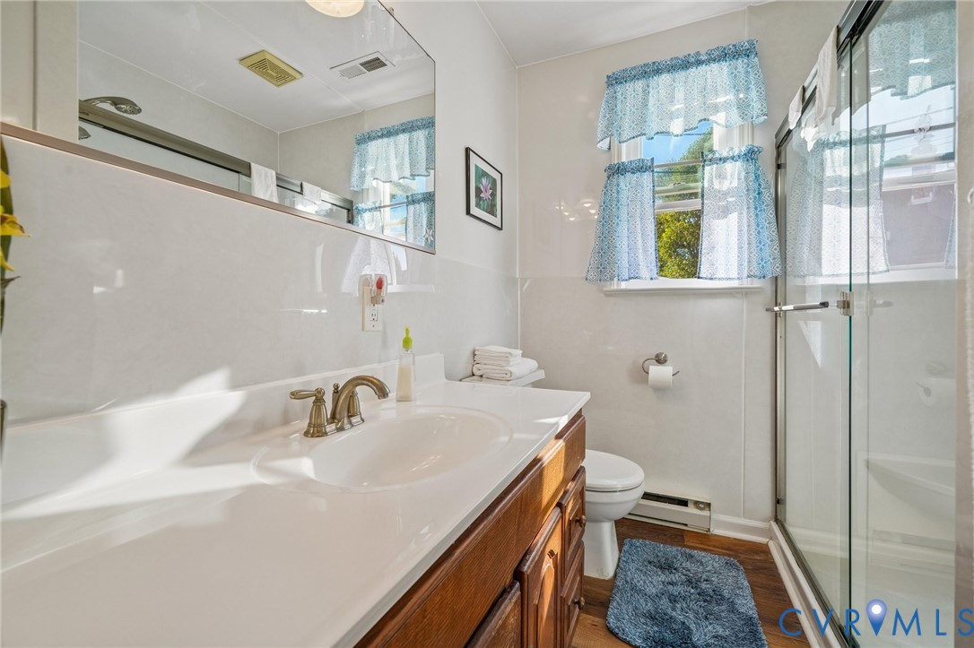 First floor Bathroom featuring a wood-finish vanity with an integrated basin, a large wall-mounted mirror, a glass-enclosed shower, and wood-finish flooring