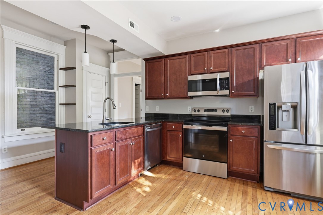 Kitchen featuring stained wood cabinetry, dark stone countertops, stainless steel appliances, wood-finish flooring, and dual pendant lighting over the peninsula