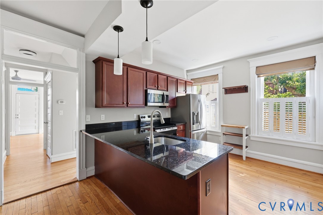 Kitchen featuring dark-toned cabinetry, a peninsula with a stone-finish countertop, stainless steel appliances, wood-finish flooring, and dual pendant lighting