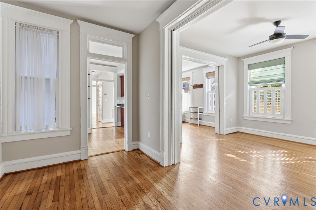 Wood-finish flooring, tall windows with heavy woodwork trim, high ceilings, transoms above interior door frames, and a ceiling fan