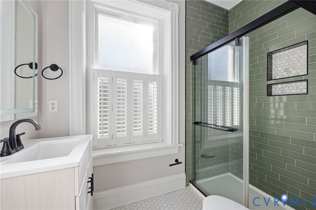 Bathroom featuring sage green subway tile shower surround with dual inset niches, glass enclosure with matte black hardware, white hexagonal tile flooring, and light wood-finish vanity