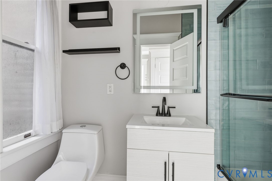 Modern vanity with an integrated sink and matte black faucet, illuminated mirror, glass-enclosed shower featuring subway-style tile, and black wall-mounted shelving