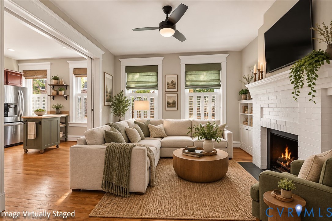 Living area featuring wood-finish flooring, a white-painted brick fireplace, built-in cabinetry, and a ceiling fan