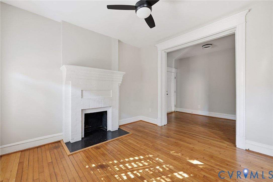 Painted brick fireplace with a dark hearth, wood-finish flooring, ceiling fan, and wide architectural cased opening
