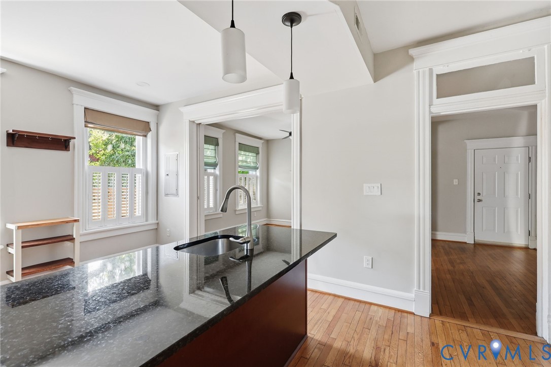 Kitchen island featuring a stone countertop, under-mount sink, and dual pendant lighting, complemented by wood-finish flooring, transom-style doorway trim, and traditional window casings