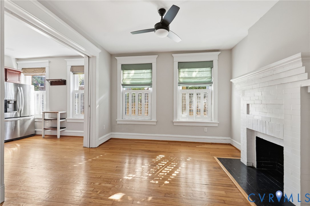 Interior space featuring wood-finish flooring, a painted brick fireplace with a contrasting hearth, multi-pane windows with traditional millwork, and a ceiling-mounted light fixture with a fan