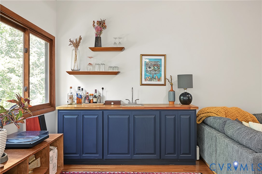 Built-in wet bar featuring navy blue shaker-style cabinetry, wood countertop, stainless steel sink basin, and two natural wood floating shelves