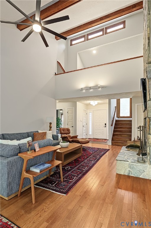 Vaulted ceilings with exposed wood beams, stone-faced fireplace, transom windows, wood-finish flooring, and multi-blade ceiling fan