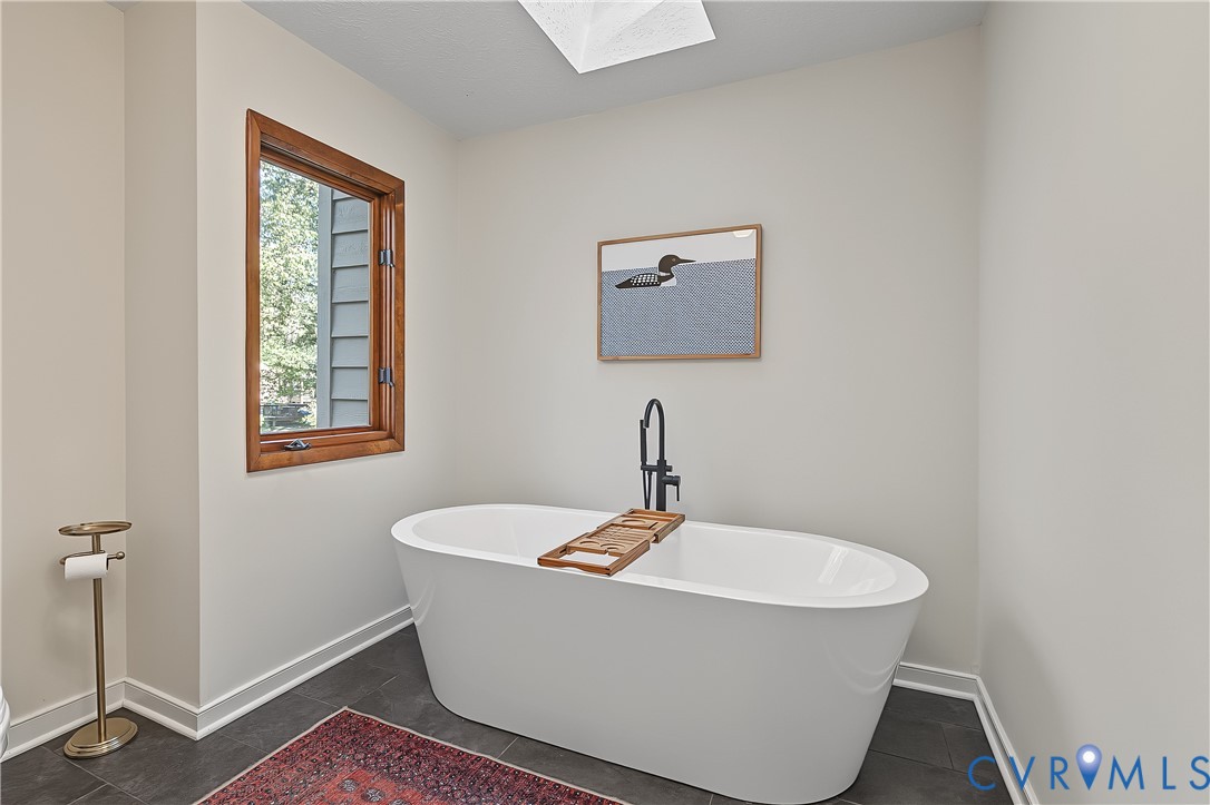 Freestanding white soaking tub with matte black floor-mounted faucet and skylight overhead, featuring dark tile flooring and wood-trimmed window