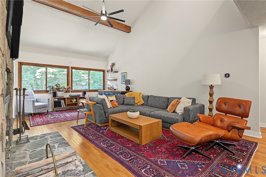 Vaulted ceiling with exposed wooden beam, floor-to-ceiling natural stone fireplace, wood-finish flooring, and ceiling fan with integrated lighting Vaulted ceiling with exposed wooden beam, floor-to-ceiling natural stone fireplace, wood-finish flooring, and ceiling fan with integrated lighting