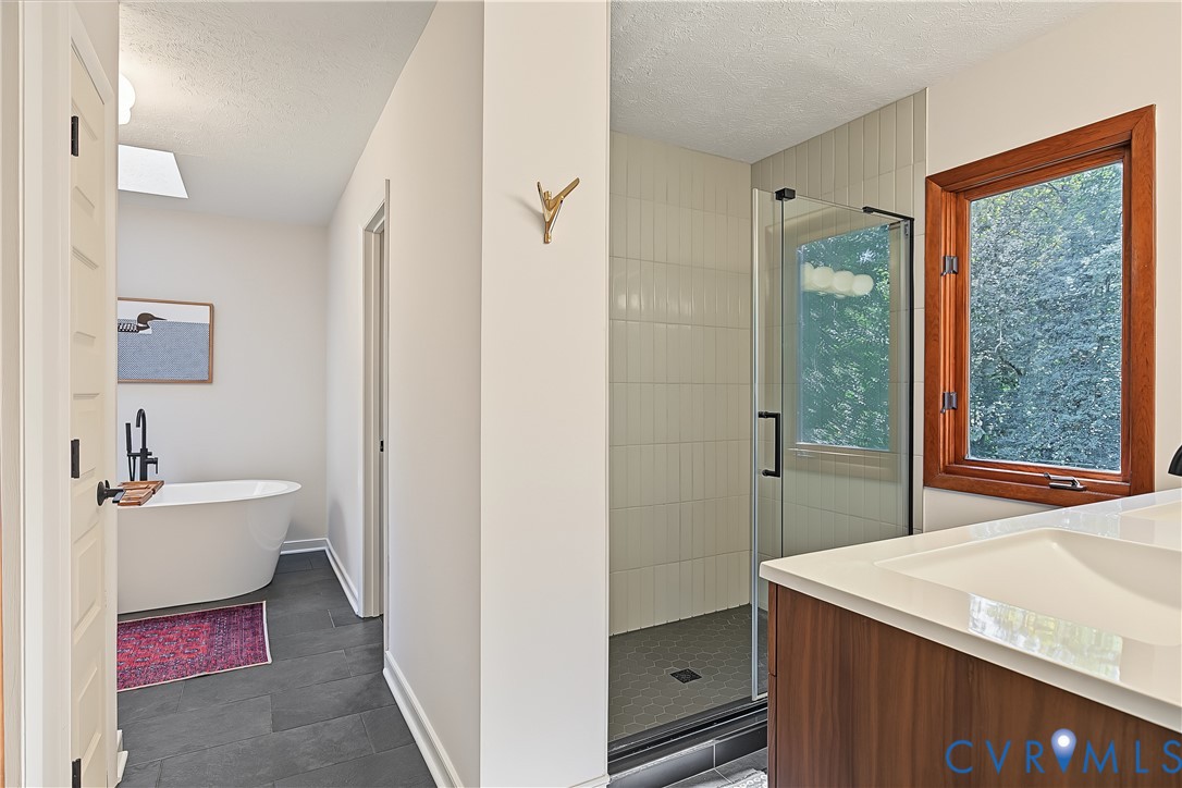 Freestanding soaking tub with matte black faucet, walk-in shower with vertical tile surround, wood-finish vanity with integrated sink, dark large-format floor tile, and wood-framed window