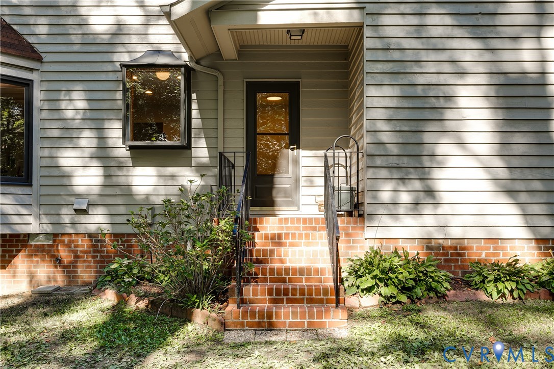 Exterior entrance featuring horizontal lap siding, brick foundation, recessed entryway, wrought iron handrails, and a multi-pane bay window
