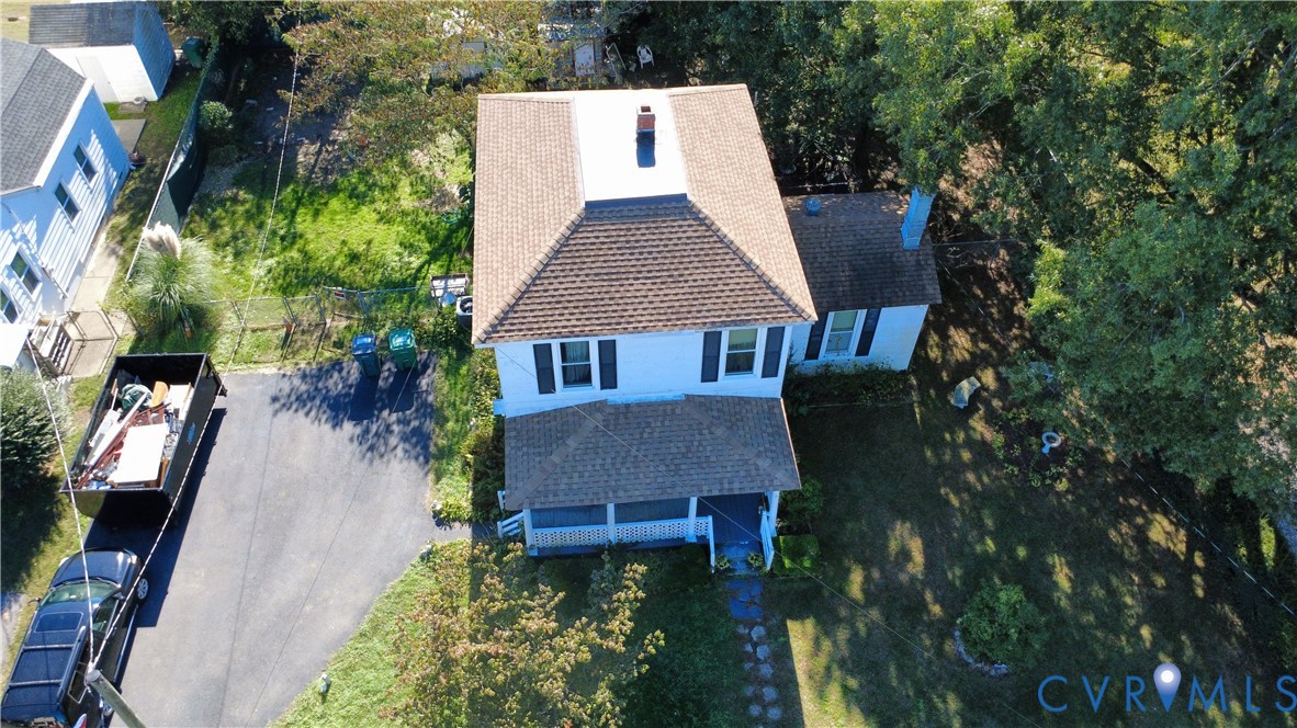 Aerial view of a detached residence featuring a shingled hipped roof, covered front porch, paved driveway, and mature landscaping on the grounds