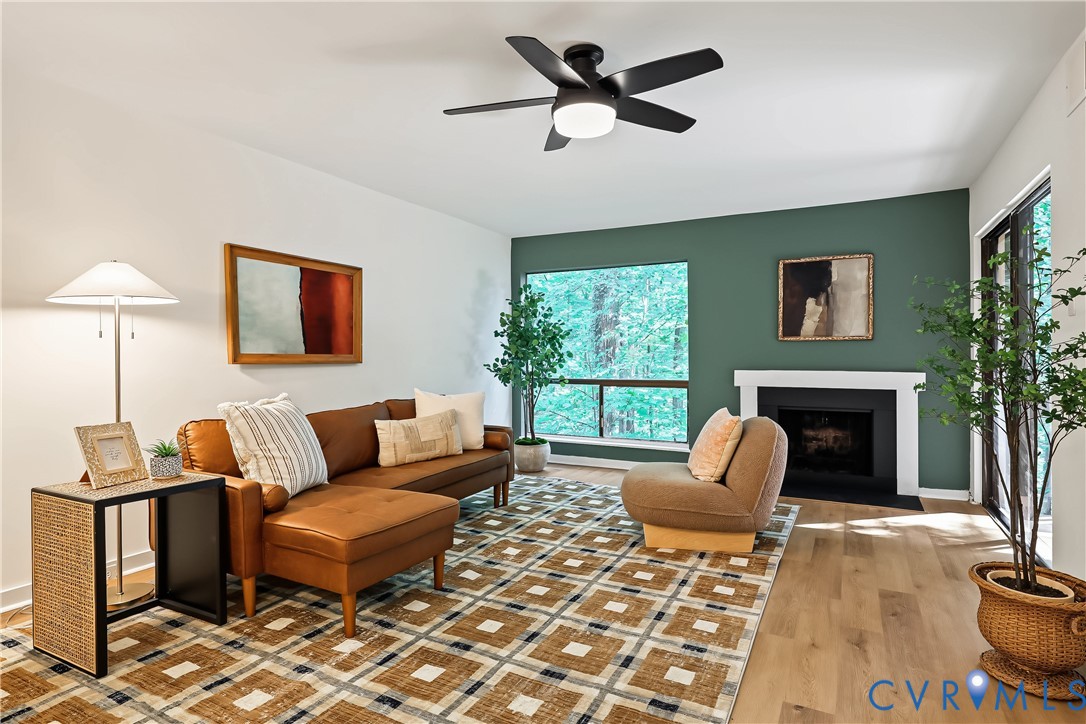 Living area featuring wood-finish flooring, a fireplace with a white mantel, an accent wall, large windows, and a ceiling fan with integrated lighting