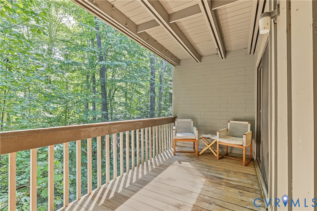 Covered balcony featuring wood decking, a wood railing with vertical balusters, exposed beam ceiling, and exterior wall-mounted lighting
