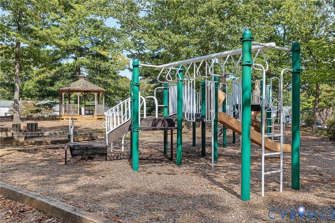 Outdoor recreational area featuring a multi-component play structure with slides and climbing apparatus, accompanied by a wooden gazebo, set within a wood-mulched landscape
