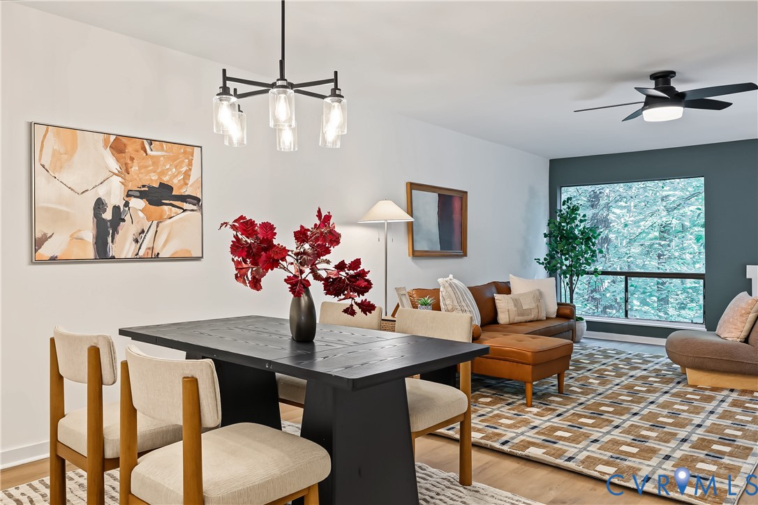 Open-concept living space featuring wood-finish flooring, a multi-light matte black chandelier, a ceiling fan with integrated lighting, and a large picture window with dark framing