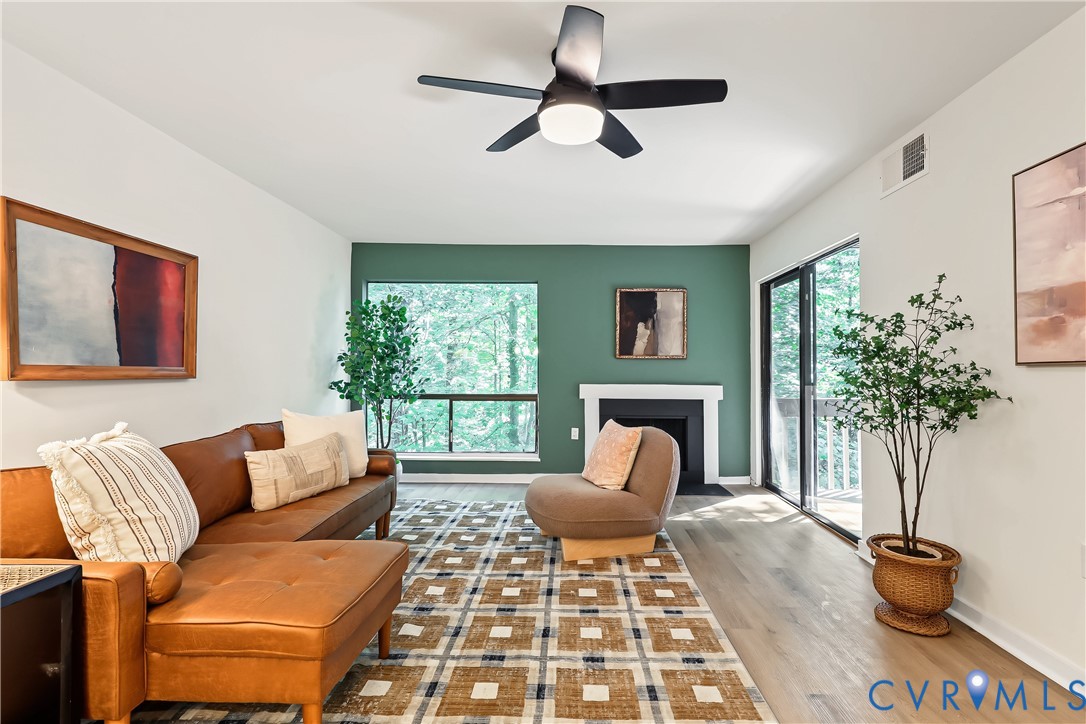 Living area featuring wood-finish flooring, accent wall, fireplace with white mantle, ceiling fan with integrated lighting, and glass sliding door