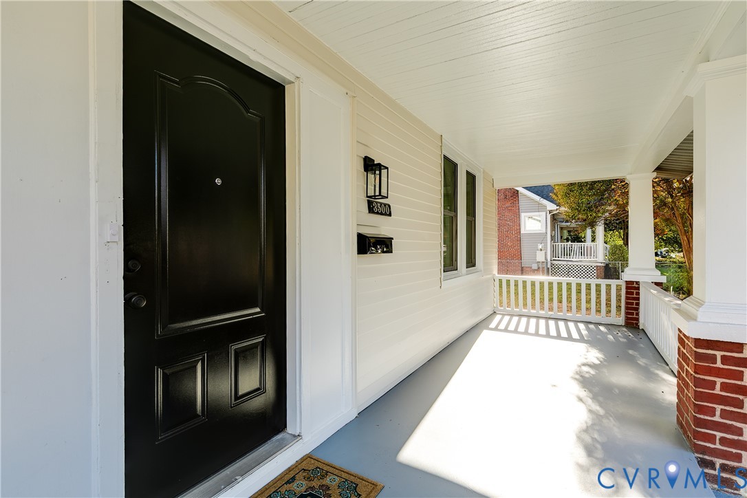 Covered front porch featuring a black panel door, beadboard ceiling, white railing with brick base columns, and wall-mounted exterior lantern lighting