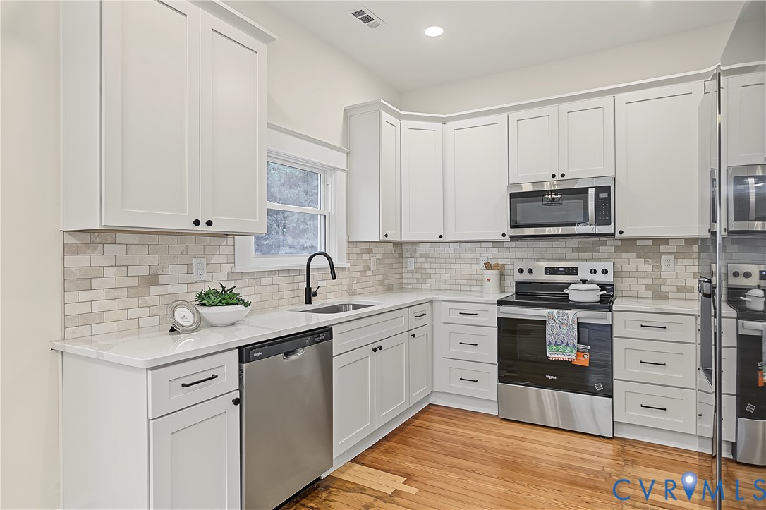 Kitchen featuring white shaker cabinetry, stone tile backsplash, stainless steel appliances, quartz countertops, and wood-finish flooring