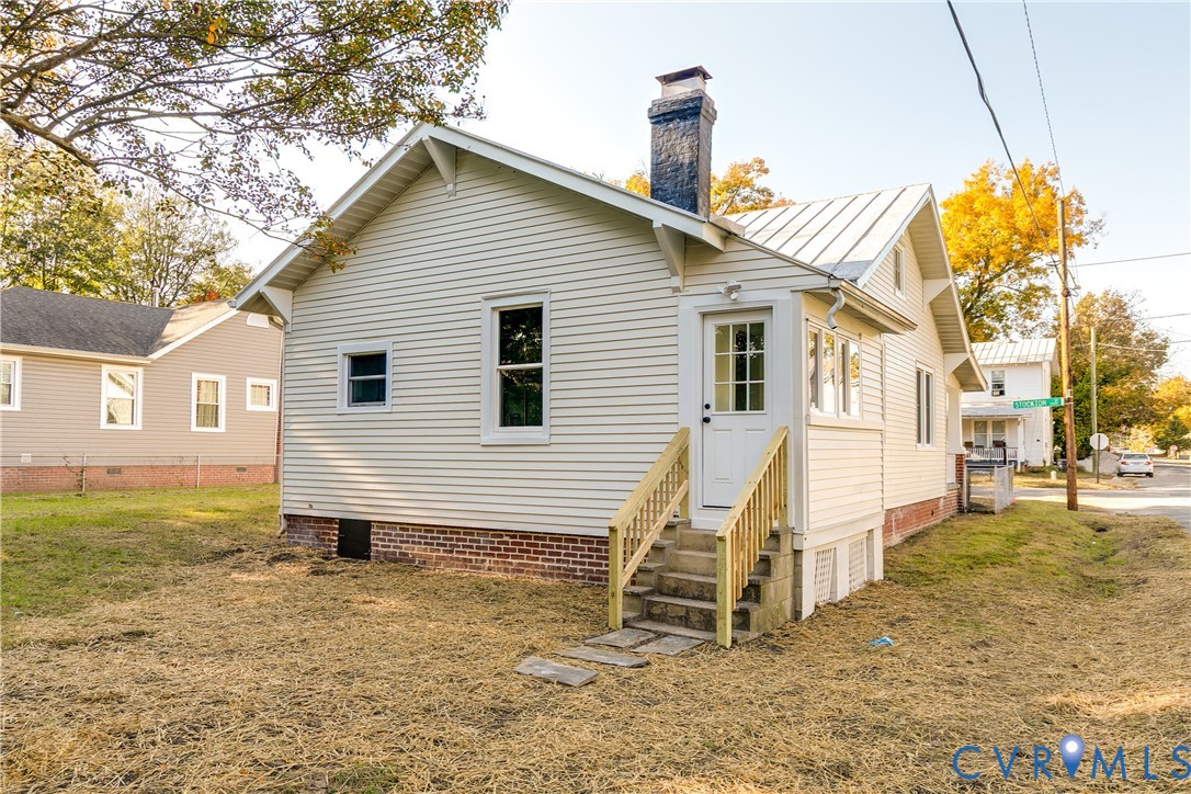 Light-colored vinyl siding, brick foundation, metal roof, exterior brick chimney, and wooden staircase with railings