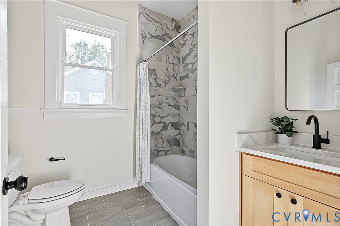 Bathroom featuring a wood-finish vanity with a stone-surfaced countertop, matte black faucet, walk-in tub with large-format patterned wall tile, and grey tile flooring