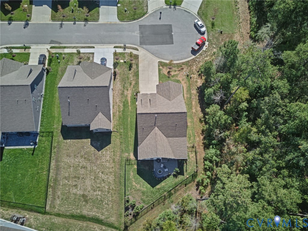 Aerial view of a residential lot featuring a concrete driveway, a multi-gabled shingle roof, a fenced backyard, and mature tree line borders