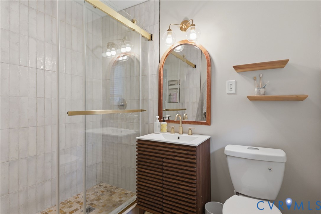Bathroom featuring a slatted wood-finish vanity, arched mirror with brass-tone frame, glass-enclosed shower with subway-style wall tile, and brass-tone plumbing fixtures