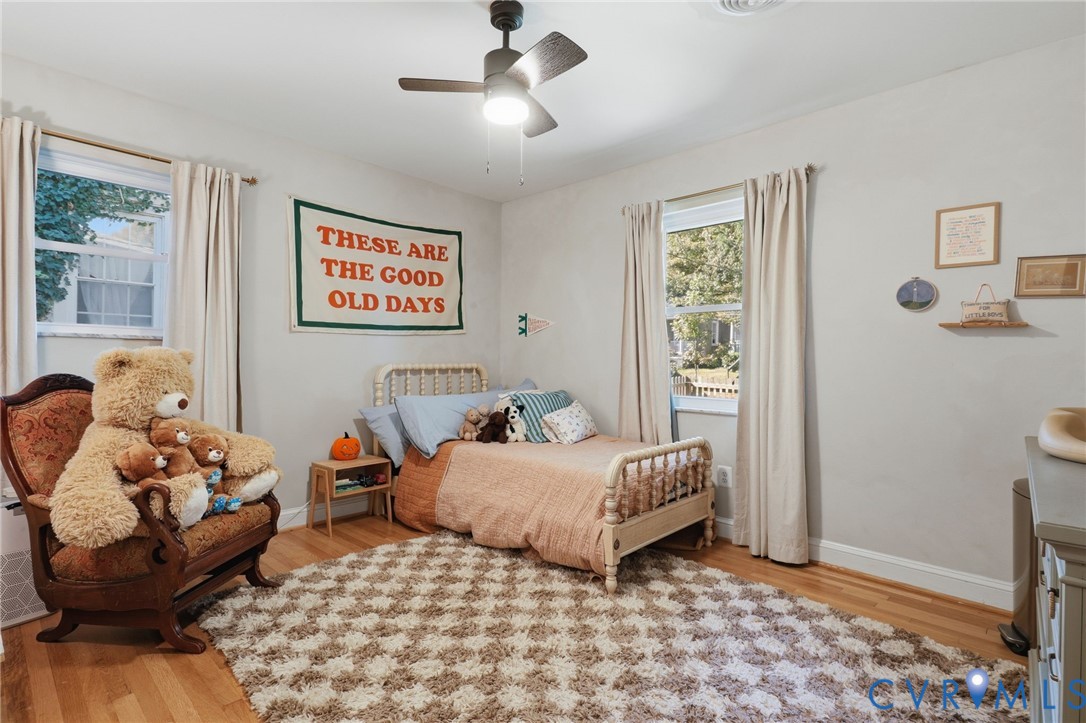 Room featuring wood-finish flooring, dual windows with natural light, and a ceiling-mounted light fixture with a multi-blade fan