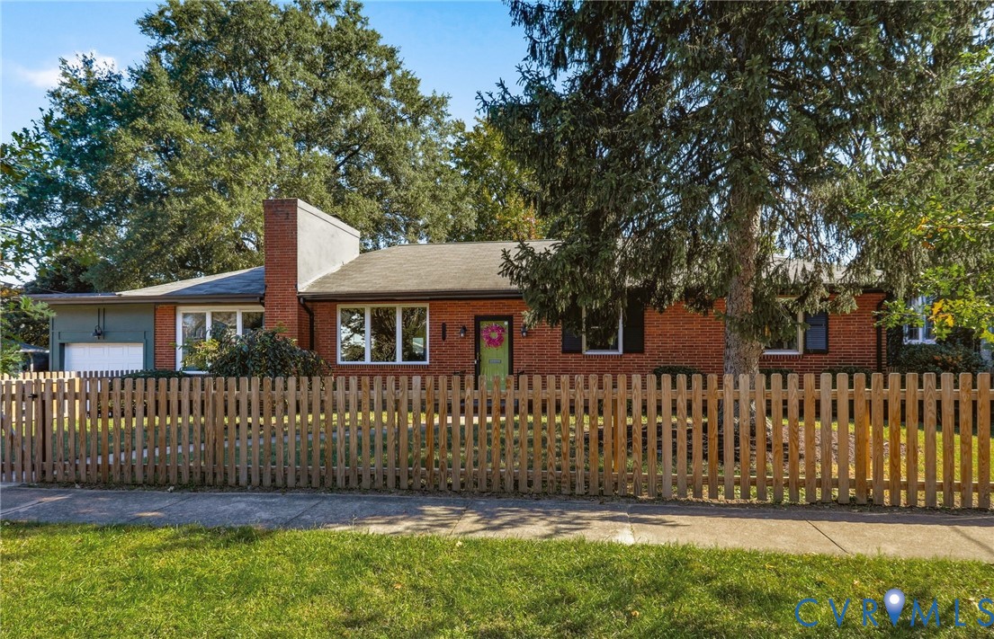 Single-story brick exterior featuring a prominent masonry chimney, attached garage, multi-pane windows, and a wood picket fence