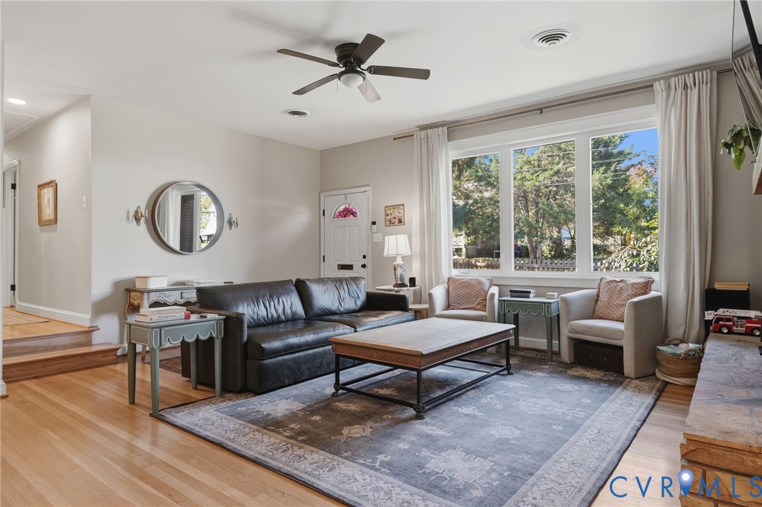 Living area featuring wood-finish flooring, a multi-pane picture window, a central ceiling fan, and a stone-clad hearth