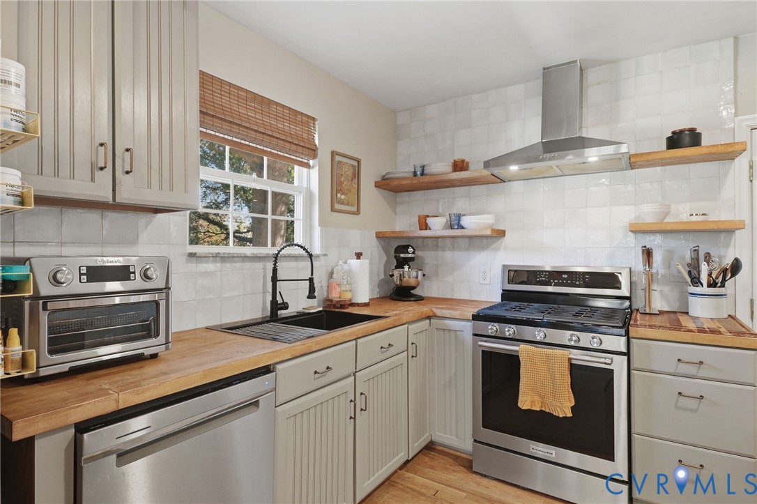 Kitchen featuring wood countertops, white tiled backsplash, stainless steel gas range with overhead ventilation, beadboard-style cabinetry, and floating wood shelving