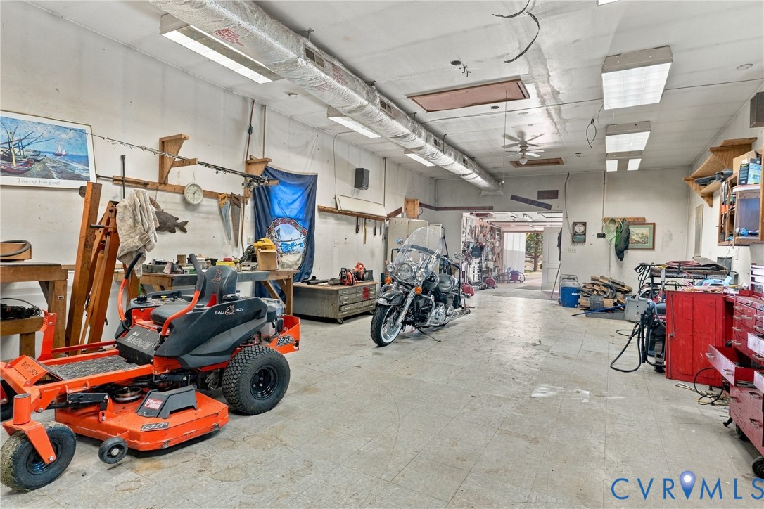 Expansive workshop space featuring concrete flooring, exposed overhead ductwork, multiple linear fluorescent light fixtures, and wall-mounted shelving units