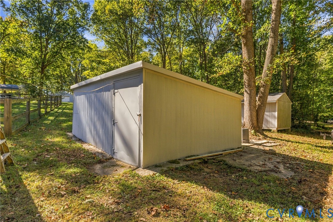 Detached storage structure with metal siding, a single-entry door, and a slanted roof, situated on a lot featuring mature trees and wood-post fencing