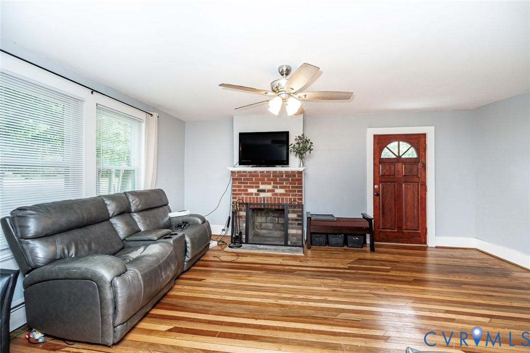 Living area featuring wood-finish flooring, a red brick fireplace with a built-in mantel, a ceiling fan with light fixture, and a solid wood exterior door with decorative glass paneling