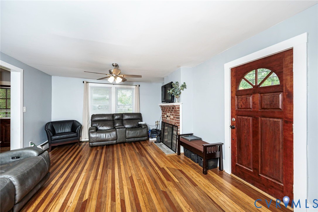 Wood-finish flooring, brick fireplace, and stained wood front door featuring an arched window detail and multipanel design, illuminated by a ceiling fan with integrated lighting