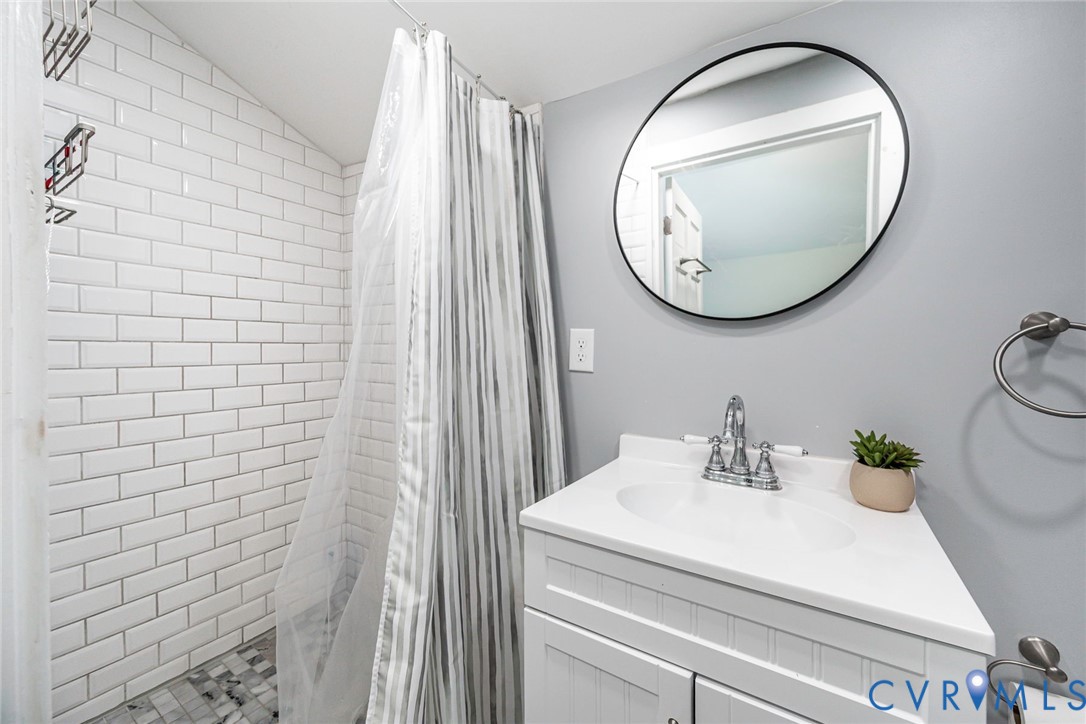 Bathroom featuring white subway tile shower surround, mosaic tile flooring, white vanity cabinet with integrated countertop, chrome dual-handle faucet, and circular wall-mounted mirror