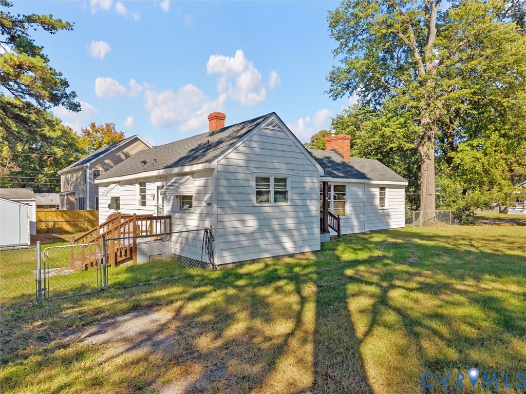 Exterior featuring white siding, two brick chimneys, a wood deck, chain-link fencing, and mature trees on a level lot