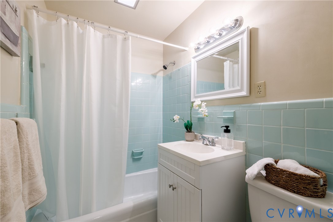 Bathroom featuring light blue wall tiles, a built-in bathtub with shower head, a white vanity cabinet with a integrated sink, and a framed wall-mounted mirror with integrated lighting